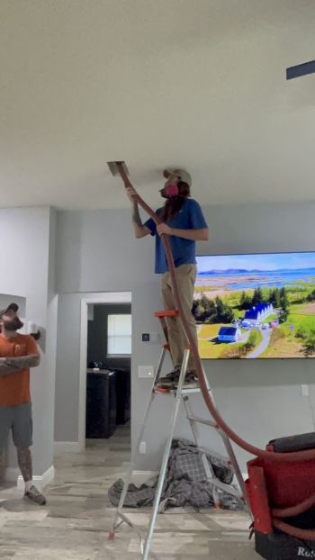 Worker on ladder installing a ceiling vent while another man watches in a modern home living room
